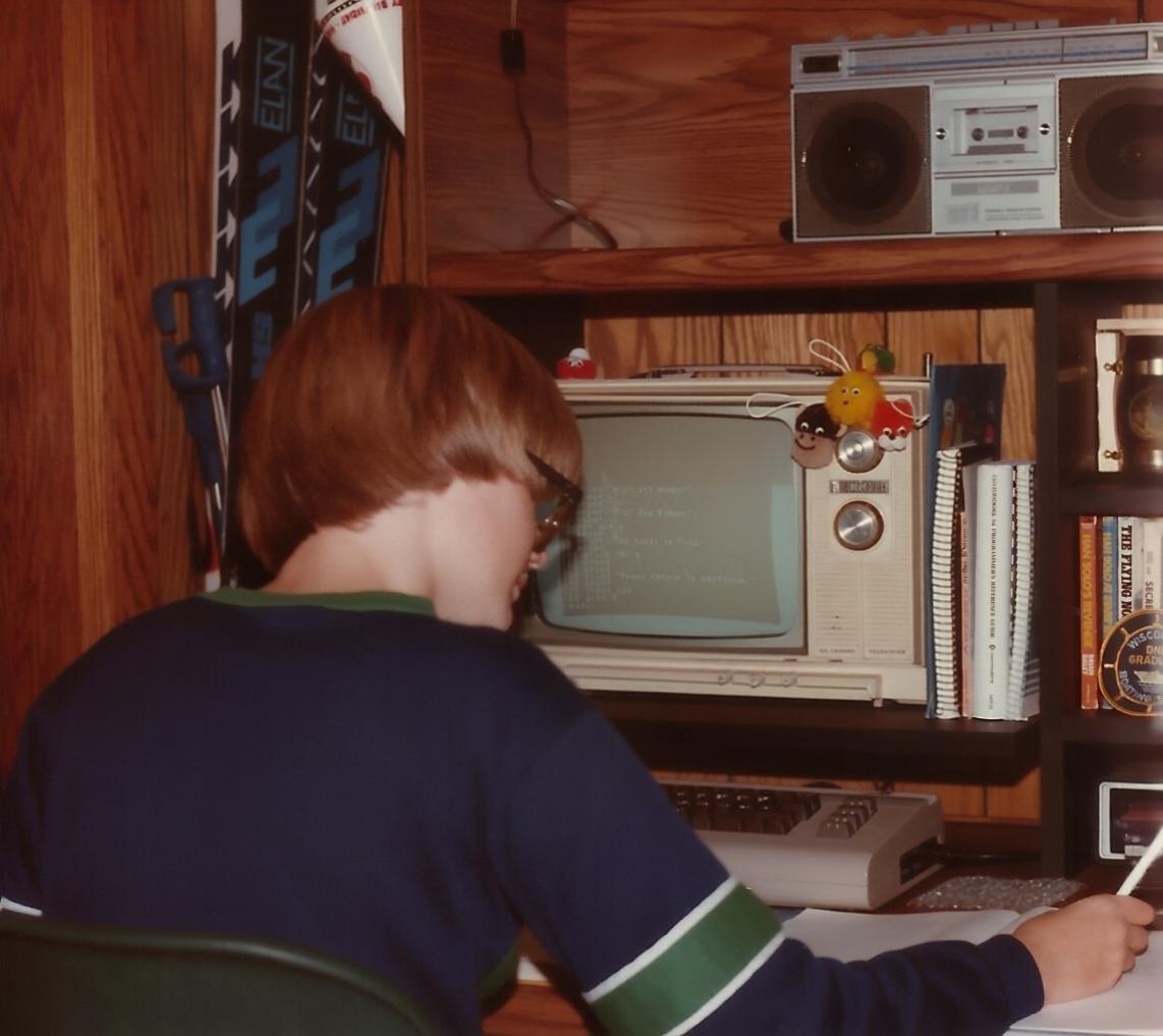 A 13 year old boy is pictured in 1983. He is sitting in a wood paneled room at a wood desk with a hutch of similar wood coloration to the walls. It is a warm colored wood. In front of him is a Commodore 64 Computer. There is a cream colored Zenith black and white TV on the desk hutch being used as a display for the computer. On the screen is BASIC code. There are various reference manuals and science fiction books on the shelves of the desk hutch. At the top shelf of the hutch is a GPX Boombox with two prominent speakers and a cassette tape deck in the middle. Two sets of downhill skis are resting against the wall next to the desk. The boy writes in a notebook. He is wearing a blue shirt with a green stripe wrapped around the arm.