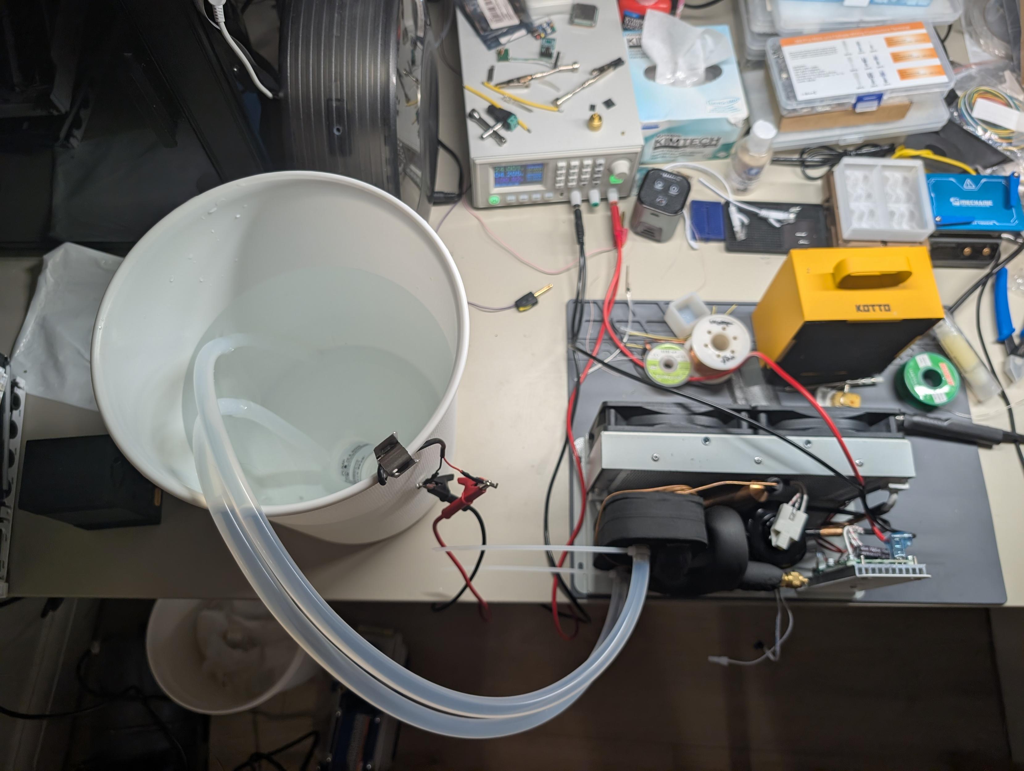 bucket of water with a pump in it and two tubes going into the heat exchanger of a vapor compression chiller. the whole thing is on the desk