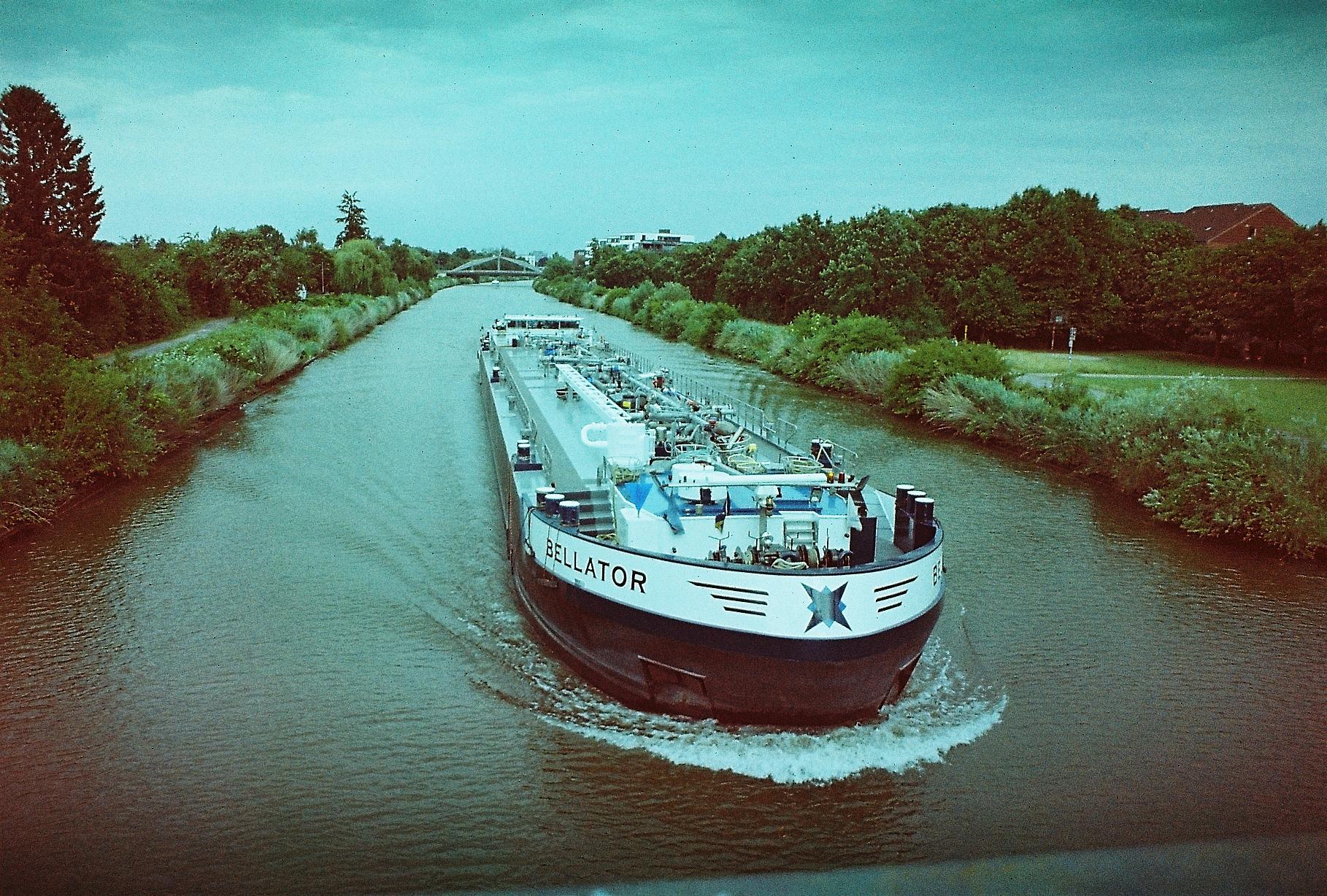 A ship going down a canal, photographed from above.
The ship's name is visible as "Bellator". The Ship is very long and slim. It is causing slight bow waves.
The scenery around the canal is green. The weather is overcast.
The colors and contrasts of the photo are off, with everything shifting slightly into blue.